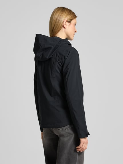 Modelka nosi czarną kurtkę przeciwdeszczową 'Regenjacke mit Omni-Tech™ ECO-Technologie' od Columbia, taliowany krój, kaptur z tunelem, kieszenie zapinane na zamek błyskawiczny, ochrona podbródka, patki przy mankietach, wydłużony tył, podszewka z siateczki, długość tyłu w rozmiarze S: 70 cm, szerokość ramion w rozmiarze S: 42 cm, szerokość w rozmiarze S: 48 cm, długość rękawów w rozmiarze S: 66 cm, widok całej sylwetki z tyłu.