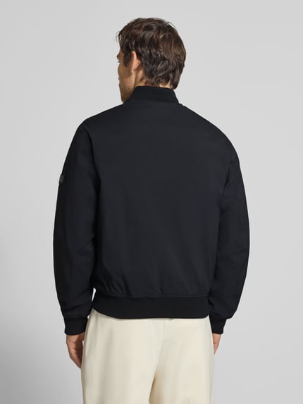 Męski model nosi bomberkę z zamkiem błyskawicznym i wpuszczanymi kieszeniami Bomberjacke mit Reißverschluss und Eingrifftaschen von JACK & JONES premium, krój regular fit, stójka, czarny materiał o gładkiej fakturze, długość tyłu w rozmiarze M: 66 cm, szerokość ramion w rozmiarze M: 42 cm, szerokość w rozmiarze M: 60 cm, długość rękawów w rozmiarze M: 68 cm, widok całej sylwetki od tyłu