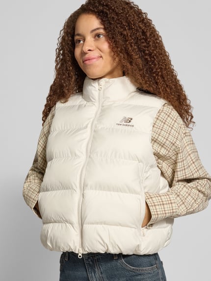 Frauenmodel trägt Steppweste mit Stehkragen Modell 'BOSTON PUFFER VEST' von NEW BALANCE, cremefarben, Struktur, Logo-Print, Reißverschlusstaschen, Ganzkörperansicht von vorne.