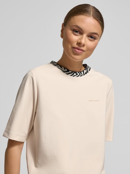 Kobiece model prezentuje damski t-shirt z mieszanki bawełny i modalu od Marc Cain, krój regular fit, logo, okrągły dekolt, zbliżenie na górną część ciała.