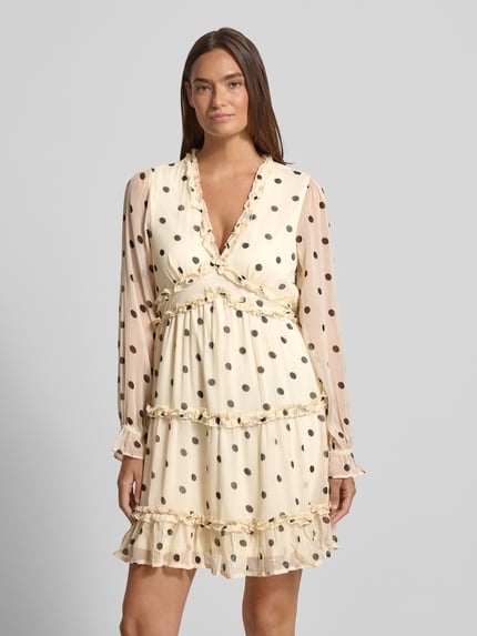 Frauenmodel trägt Regular Fit Sommerkleid aus Chiffon Modell 'SMILLA' von Vero Moda in Optik:Allover-Muster, Mini-Länge, V-Ausschnitt, Rüschen-Applikation, Ballonärmel, langarm, ohne Verschluss, Materialverarbeitung: Chiffon, helle Grundfarbe mit schwarzen Punkten, zarte und leichte Textur, Ganzkörperansicht von vorne.