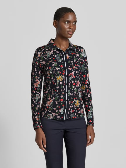 Modelka nosi Slim Fit Hemdbluse mit floralem Allover-Print von Marc Cain, elastyczna mieszanka materiałów, krój slim fit, kwiatowy nadruk na całej powierzchni, kołnierzyk koszulowy, bluzka z długim rękawem, długość tyłu w rozmiarze 36: 59 cm, długość rękawów w rozmiarze 36: 63 cm, kolorowe wzory na ciemnym tle, jednolite spodnie, gładka faktura, Ganzkörperansicht z przodu.