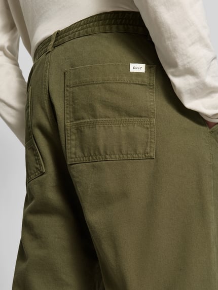 Männermodel trägt Stoffhose mit Label-Patch von Forét aus reiner Baumwolle in Olivgrün, strukturiertes Gewebe, Detailaufnahme, KI-modifiziertes Bild.