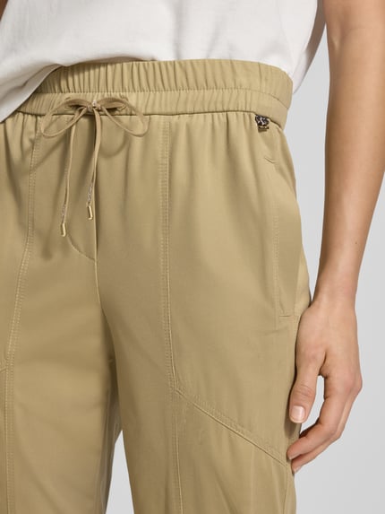 Frauenmodel trägt Slim Fit Stoffhose mit Reißverschlusstaschen von Marc Cain in Beige, elastischer Bund mit Tunnelzug, Label-Details, Nahaufnahme Oberkörper