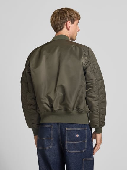 Männermodel trägt Bomberjacke mit Pattentaschen und Label-Applikation von Alpha Industries in Olivgrün, glatte Textur, Ganzkörperansicht Rückansicht, Regular Fit, Stehkragen, Reißverschluss, Kontext: Rückenlänge 69 cm, Schulterbreite 44 cm, Brustweite 54 cm, Ärmellänge 64 cm.