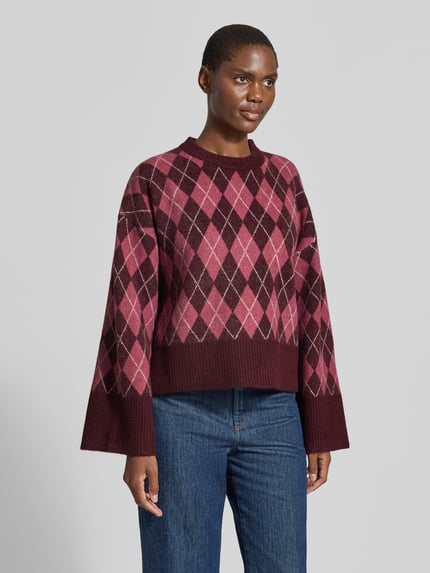Kobiece model nosi Relaxed Fit Pullover mit Woll-Anteil od Neo Noir, elastyczna mieszanka materiałów, imitacja dzianiny, okrągły dekolt, długość tyłu w rozmiarze 36: 54 cm, długość rękawów w rozmiarze 36: 50 cm, bordowe i różowe romby, widok całej sylwetki z przodu.