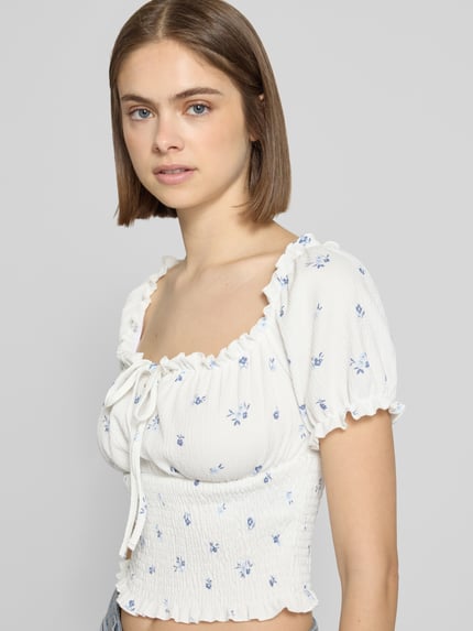 Frauenmodel trägt Slim Fit Blusentop mit Allover-Muster Modell 'HILLARY' von Only, weiß-blau, Struktur mit floralem Muster, Henley-Ausschnitt, Kurzarm, Cropped-Länge, Raffungen am Ärmelabschluss, Ganzkörperansicht von vorne.