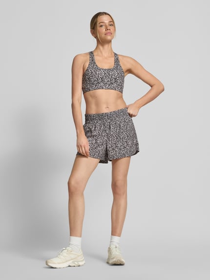 Frauenmodel trägt Running Shorts mit elastischem Bund Modell 'kallin' von VARLEY im Damen-Relaxed-Fit aus Material-Mix mit Stretch-Anteil, grafisches Allover-Muster mit kurzen Beinen, Bundweite bei Größe S 40 cm, Innenbeinlänge 8 cm. Ganzkörperansicht von vorne.