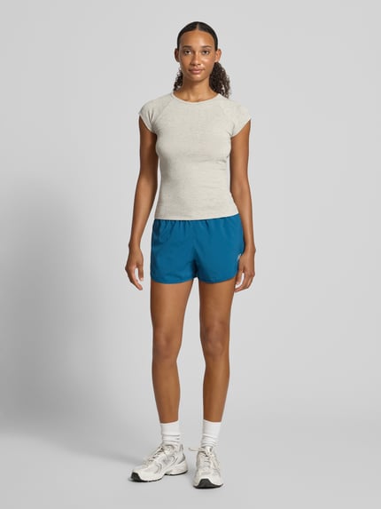 Frauenmodel trägt blaue Shorts mit Label-Print Modell 'ESSENTIAL' von New Balance, elastischer Bund, schlichter Hintergrund, Ganzkörperansicht von vorne.