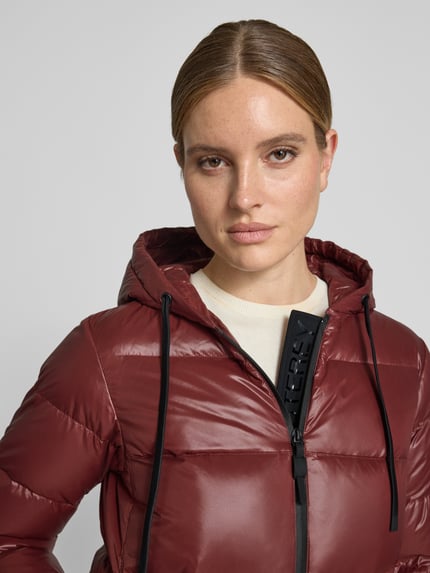 Frauenmodel trägt Steppjacke mit Kapuze und Eingrifftaschen Modell 'KENID' von Peuterey in Dunkelrot, glatte Oberfläche, Nahaufnahme Oberkörper. Gerader Schnitt, Reißverschluss, sichtbarer Logo-Detail, Größe 36: Rückenlänge 61 cm, Schulterbreite 41 cm, Brustweite 48 cm, Ärmellänge 60 cm.