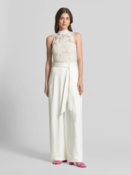 Frauenmodel trägt Jumpsuit mit Häkelspitze Modell 'CALANTA' von Ted Baker aus weißem Baumwoll-Mix, Taillierter Schnitt, Bindegürtel, Seitliche Eingrifftaschen, gerippter Rundhalsausschnitt, Ganzkörperansicht von vorne.