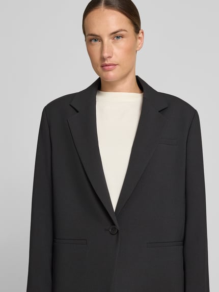 Kobiece model nosi żakiet Relaxed Fit Blazer mit Paspeltaschen Modell 'MOWTIE' od Drykorn, mieszanka materiałów z dodatkiem wiskozy, kołnierz z połami, zapięcie na guzik, kieszenie z wypustkami, długość tyłu w rozmiarze 36: 72 cm, szerokość w rozmiarze 36: 48 cm, długość rękawów w rozmiarze 36: 60 cm, kolor czarny, gładka faktura, widok z przodu, zbliżenie na górną część ciała.