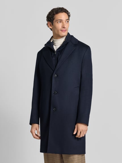 Männermodel trägt Mantel aus Wolle mit integriertem Stehkragen von BOSS in dunkelblau, unifarben mit Struktur, seitliche Taschen, Ganzkörperansicht von vorne.
