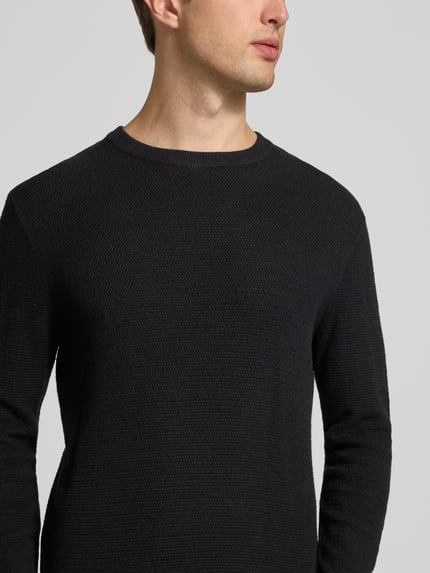 Męski model ma na sobie sweter z dzianiny z bawełny, Model 'Ryder' od JACK & JONES, krój slim fit, imitacja dzianiny, czarny, okrągły dekolt, prążkowane wykończenia, logo, długie rękawy, widok na górną część ciała z przodu.