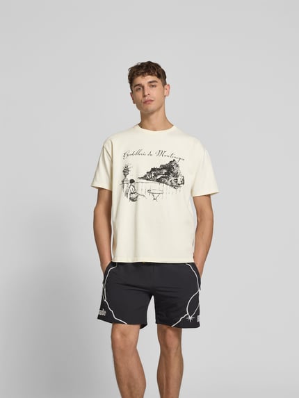 Mannelijk model draagt Regular Fit T-shirt met motiefprint van Rhude in crème met zwarte print, puur katoen, ronde hals, ruglengte maat M 66 cm, geheellichaamsaanzicht van voren.