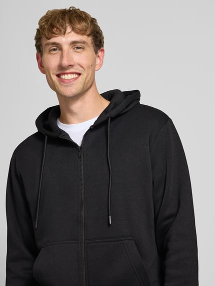 Męski model nosi Regular Fit Sweatjacke aus Baumwoll-Mix Modell 'BRADLEY' od JACK & JONES, czarny kolor, miękka faktura, kaptur z tunelikiem, kangurka i elastyczne wykończenia, Długość tyłu w rozmiarze M: 67 cm, Długość rękawów w rozmiarze M: 68 cm, mieszanka bawełny, zbliżenie na górną część ciała.