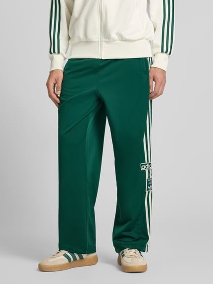Mężczyzna nosi spodnie dresowe model "ADIBREAK" od adidas Originals, luźny krój, elastyczny pas, wpuszczane kieszenie, wyhaftowane logo, długość wewnętrzna nogawki w rozmiarze M: 74 cm, zielony odcień, gładka tekstura, całkowity widok z przodu.