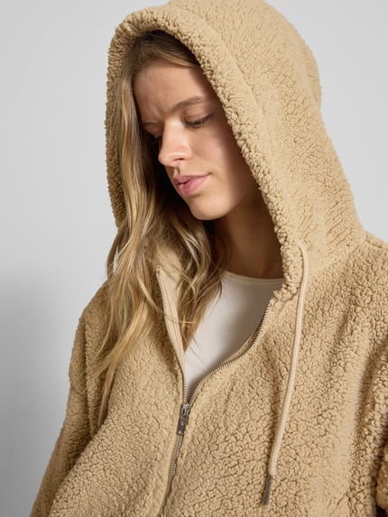 Kobiece model nosi Kurtka z futerka o luźnym, krótkim kroju, model 'CUDDLE' od Noisy May z kapturem, zamkiem i wpuszczanymi kieszeniami, beżowa faktura, detal: rozmiar S dł. 48 cm, szer. ramion 79 cm, szer. klatki 77 cm, dł. rękawów 48 cm, zbliżenie na górną część ciała.