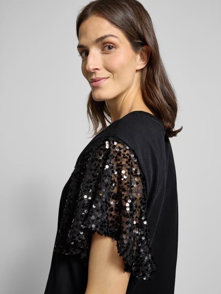 Frauenmodel trägt T-Shirt mit geripptem Rundhalsausschnitt Modell 'WINGS' von FREE/QUENT aus schwarzer Baumwolle, Paillettenbesatz am Ärmel, Nahaufnahme Oberkörper.