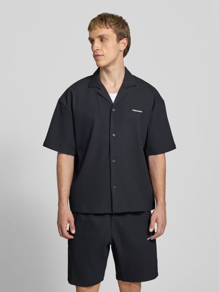 Männermodel trägt Relaxed Fit Freizeithemd mit Reverskragen Modell 'Troy' von Pegador, Schwarz, Strukturmuster, Ganzkörperansicht von vorne. Rückenlänge bei Größe M: 67 cm, Schulterbreite: 49 cm, Ärmellänge: 24 cm, durchgängige Knopfleiste, kurzärmelig, Logo-Stitching.