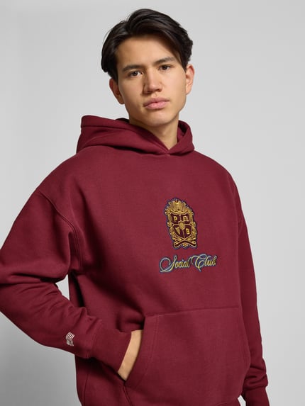 Männermodel trägt Hoodie mit Kapuze von PROHIBITED in Dunkelrot, Baumwoll-Mix, Label-Detail, Känguru-Tasche, Nahaufnahme Oberkörper.
