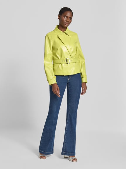 Frauenmodel trägt Jacke mit Gürtel und Ärmelriegeln von Marc Cain in Gelb, sichtbare Struktur, kombiniert mit blauer Jeans, Ganzkörperansicht von vorne, Regular Fit, Hakenverschluss, Rückenlänge 58 cm, Schulterbreite 40 cm, Brustweite 48 cm, Ärmellänge 61 cm.