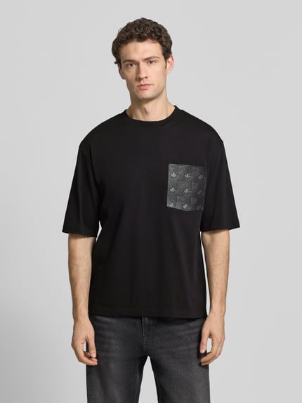Männermodel trägt Loose Fit T-Shirt aus reiner Baumwolle von Dsquared2, Schwarz mit Leder-Brusttasche und Logo, Ganzkörperansicht von vorne.