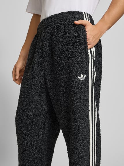 Mannenmodel draagt Sweatpants mit elastischem Bund van adidas Originals in bouclé-look, losse pasvorm, zwart met witte strepen, detailopname heup.