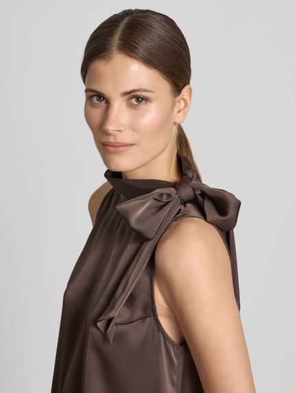 Kobiece modelka nosi Blusentop aus Satin mit Spitzenbesatz Modell 'LORENZ' od Neo Noir, kolor brązowy, kokarda, krój regular fit, stójka, obszycie koronką, satynowa powierzchnia, długość tyłu 65 cm, długość rękawów 6 cm, zbliżenie na górną część ciała.