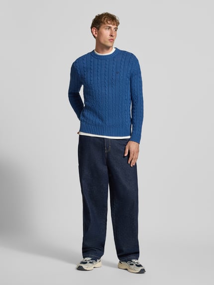 Männermodel trägt Strickpullover mit Zopfmuster und Rundhalsausschnitt von Superdry in Blau, Struktur und Slim Fit, Ganzkörperansicht von vorne.