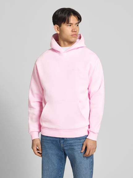Męski model trägt Hoodie mit Känguru-Tasche von REVIEW aus mieszanka bawełny, luźny krój, kaptur, detal z logo, kieszeń kangurka, elastyczne wykończenia, długość tyłu w rozmiarze M: 68 cm, długość rękawów w rozmiarze M: 62 cm; pastelowy róż, gładka faktura, niebieskie spodnie, Ganzkörperansicht von vorne
