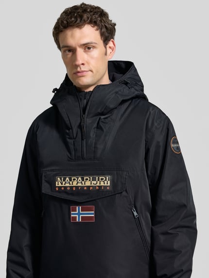 Männermodel trägt Jacke mit Kapuze und kurzem Reißverschluss von Napapijri in Schwarz mit Label-Patches, Nahaufnahme Oberkörper. Modell RAINFOREST NEXT, gerader Schnitt, robuste Textur.