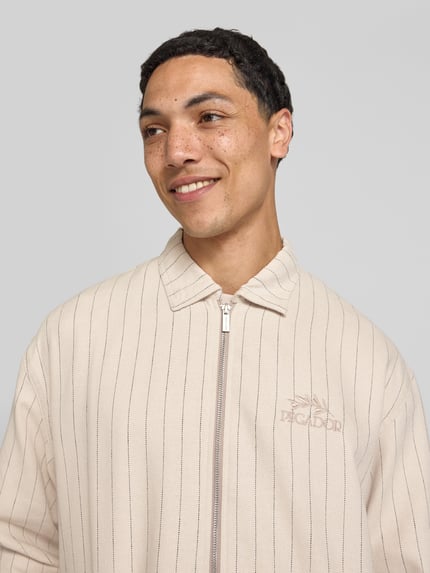 Mężczyznamodel nosi Overshirt aus Viskose-Mix mit Logo-Stitching Modell 'Sistelo' von Pegador, mieszanka wiskozy, krój relaxed fit, kołnierzyk typu kent, zamek błyskawiczny, wyhaftowane logo, kolor beżowy w prążki, długość tyłu w rozmiarze M: 69 cm, Nahaufnahme górnej części ciała.