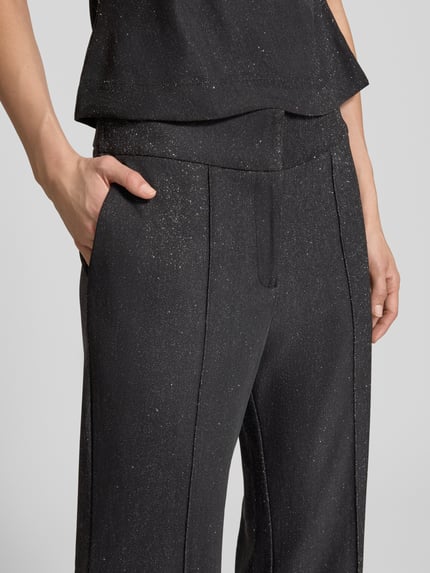 Kobieta nosi Flared Stoffhose mit Glitter-Effekt od YAS, czarne spodnie materiałowe z efektem błyszczącym, zakładki w pasie, wpuszczane kieszenie, elastyczny pas, zbliżenie na obszar talii i kieszeni.