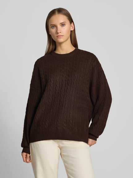Frauenmodel trägt Oversized Fit Strickpullover mit Zopfmuster von REVIEW in Dunkelbraun, Struktur mit elastischen Abschlüssen, Ganzkörperansicht von vorne. Rückenlänge bei Größe S: 64 cm, Ärmellänge 50 cm, elastischer Material-Mix.