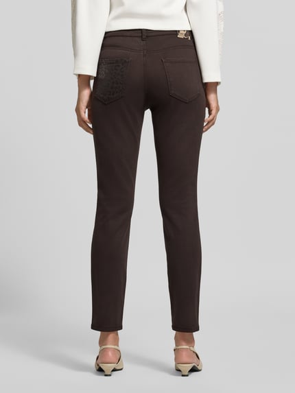 Kobieta nosi Slim Fit Jeans mit Lyocell-Anteil von Marc Cain, elastyczna mieszanka materiałów, krój slim fit, 5 kieszeni, Długość 7/8, Szerokość w rozmiarze 36: 36 cm, Długość wewnętrzna nogawki w rozmiarze 36: 72 cm, ciemnobrązowy kolor, gładka tekstura, widok całej sylwetki od tyłu.