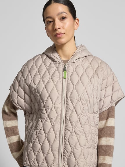 Modelka kobieca nosi Steppjacke im kurzärmeligen Design od Fuchs Schmitt w beżowym kolorze z kapturem i krótkimi rękawami, luźny krój, zapięcie na zamek, kieszenie boczne, długość tyłu w rozmiarze 36: 65 cm, szerokość ramion w rozmiarze 36: 47 cm, szerokość klatki piersiowej w rozmiarze 36: 57 cm, tekstura pikowana, z przodu, zbliżenie na górną część ciała