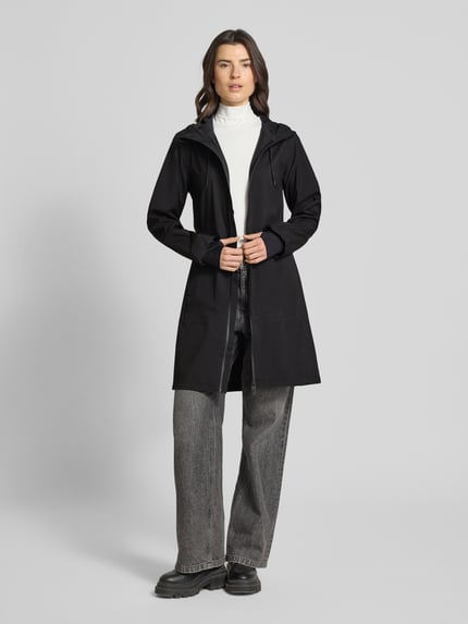 Frauenmodel trägt Regular Fit Mantel Daumenschlaufen Modell 'Suva' von Rains, czarny, krój regular fit, kaptur z tunelem, zamek błyskawiczny dwustronny, taliowany, wpuszczane kieszenie z zamkiem, długość tyłu w rozmiarze S: 94 cm, szerokość ramion w rozmiarze S: 38 cm, szerokość klatki piersiowej w rozmiarze S: 45 cm, długość rękawów w rozmiarze S: 61 cm, całkowity widok z przodu.