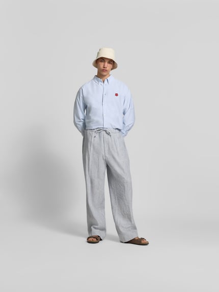Mężczyzna nosi Wide Fit Stoffhose aus Leinen-Mix od Officine Générale, biało-szare paski, luźny krój, widok całej sylwetki z przodu.