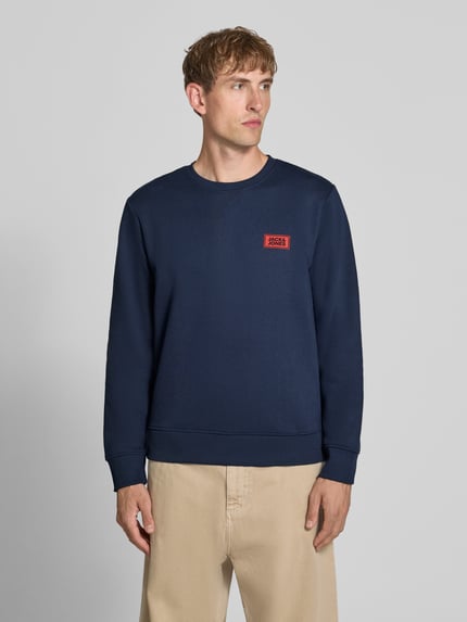 Męski model nosi bluzę z mieszanki bawełny o luźnym kroju, okrągły dekolt, nadruk z logo, prążkowane wykończenia, Model 'RUSH' od JACK & JONES, tył 67 cm, rękawy 67 cm, ujęcie całej sylwetki z przodu