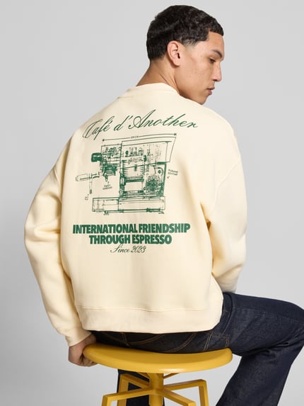 Męski model nosi Regular Fit Sweatshirt mit Logo-Print od ANOTHER COTTON LAB, mieszanka bawełny, okrągły dekolt, elastyczne wykończenia, długość tyłu w rozmiarze M: 63 cm, długość rękawów: 58 cm, kremowa bluza z zielonym nadrukiem i teksturą, widok na cały tył.