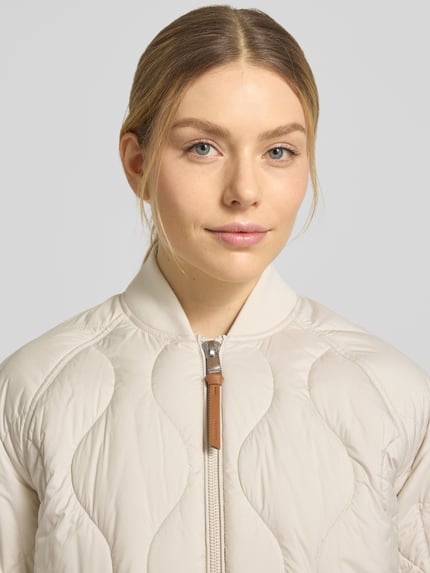 Frauenmodel trägt Steppmantel mit Ärmeltasche Modell 'AVON' von Beaumont Amsterdam in Beige, gesteppte Struktur, Nahaufnahme Oberkörper, gerade Passform, Stehkragen, Zweiwege-Reißverschluss.