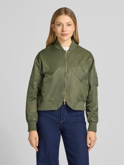 Frauenmodel trägt Bomberjacke mit Zweiwege-Reißverschluss von Original Bombers in Olivgrün mit Stehkragen und Stitching, Ganzkörperansicht von vorne.