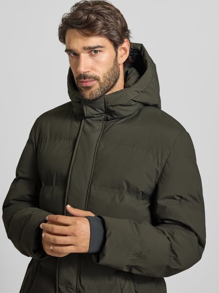 Männermodel trägt Steppmantel mit verdecktem Reißverschluss und Eingrifftaschen von MCNEAL, olivgrün, matte Oberfläche, Nahaufnahme Oberkörper. Rückenlänge 100 cm, Schulterbreite 48 cm, Brustweite 59 cm, Ärmellänge 64 cm, Regular Fit, Kapuze.