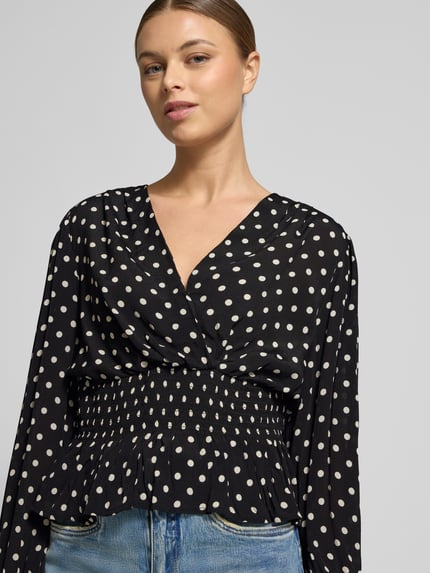 Vrouwenmodel draagt Blouse uit zuivere viscose in wikkellook Model 'Tamona' van Neo Noir, regular fit, met ruches, V-hals, lange mouwen, getailleerd, zwarte stof met witte stippen, ruglengte maat 36: 58 cm, mouwlengte: 66 cm, close-up bovenlichaam.