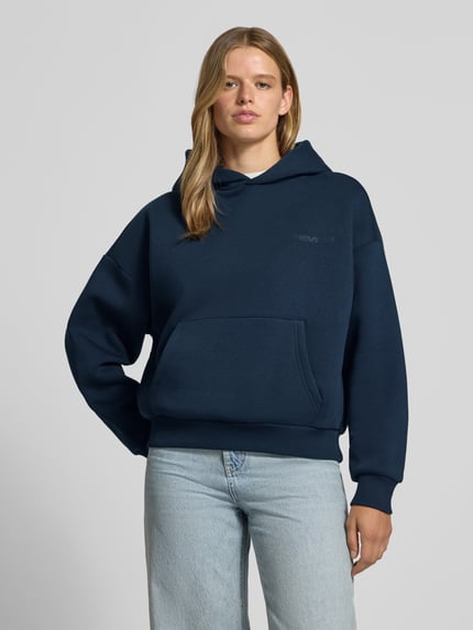 Frauenmodel trägt Kapuzenpullover aus Baumwoll-Mix von REVIEW in Dunkelblau mit Känguru-Tasche, lockerer Passform und gerippten Bündchen, Ganzkörperansicht von vorne.