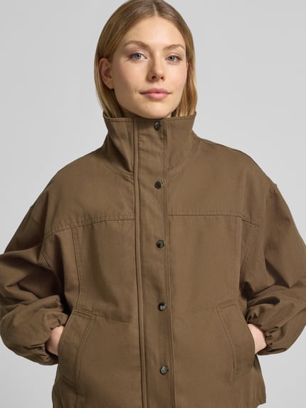 Frauenmodel trägt Bomberjacke mit Stehkragen Modell 'Selio' von Neo Noir, Material-Mix mit Stretch-Anteil, Lockerer-Schnitt, Seitliche Eingrifftaschen, Reiß- und Druckknopfverschluss, elastische Ärmelabschlüsse, Farbe Braun, mittleres Braun, matte Textur, Ganzkörperansicht von vorne. Rückenlänge 55 cm, Schulterbreite 49 cm, Brustweite 48 cm, Ärmellänge 51 cm bei Größe 36.