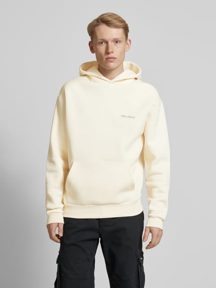 Männermodel trägt Hoodie mit Label-Print von REVIEW in Beige, gerader Schnitt, Känguru-Tasche, elastische Abschlüsse, Ganzkörperansicht von vorne. Baumwoll-Mix, Kapuze, Rückenlänge 68 cm, Ärmellänge 67 cm.