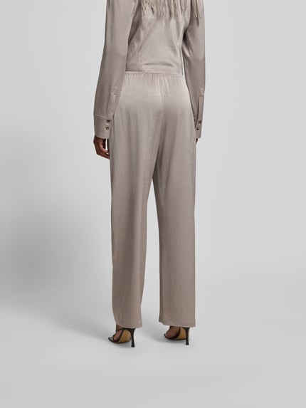 Frauenmodel trägt Stoffhose mit elastischem Bund von Nanushka aus Samt in Beige, relaxed Fit, Ziernähte, Beinabschlüsse, Ganzkörperansicht Rückansicht.