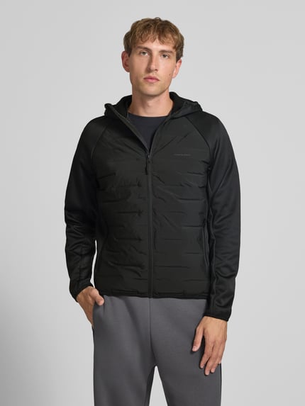 Männermodel trägt Hybrid Jacke mit Kapuze Modell 'PERFECT' von JACK & JONES in Schwarz mit Reißverschluss und Logo-Print, Ganzkörperansicht von vorne. Gerader Schnitt, seitliche Reißverschlusstaschen, glatte und gesteppte Texturen, Kontext: Herren Hybrid Jacke, Rückenlänge 67 cm, Schulterbreite 44 cm, Brustweite 51 cm, Ärmellänge 69 cm.